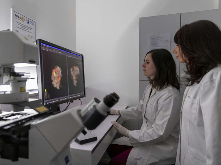Deux chercheuses avec blouse et gants regardent un écran derrière un microscope. On y voit des cellules en gros plans