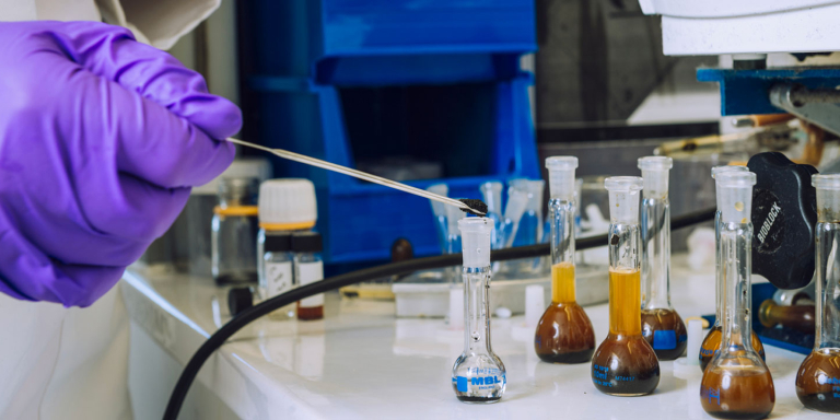 Au premier plan, une main recouverte d'un gant violet manipule des fioles